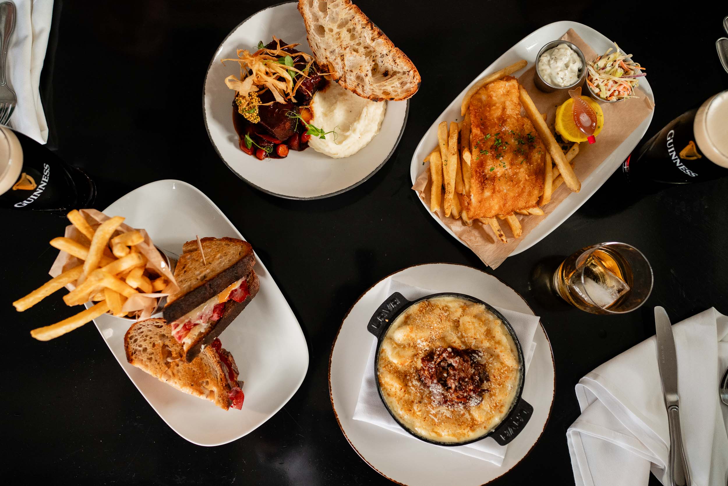 four dishes from an aerial perspective including beer battered haddock over fries, Corned Beef & Cabbage Sandwich, short rib with mashed potatoes, and mac & cheese