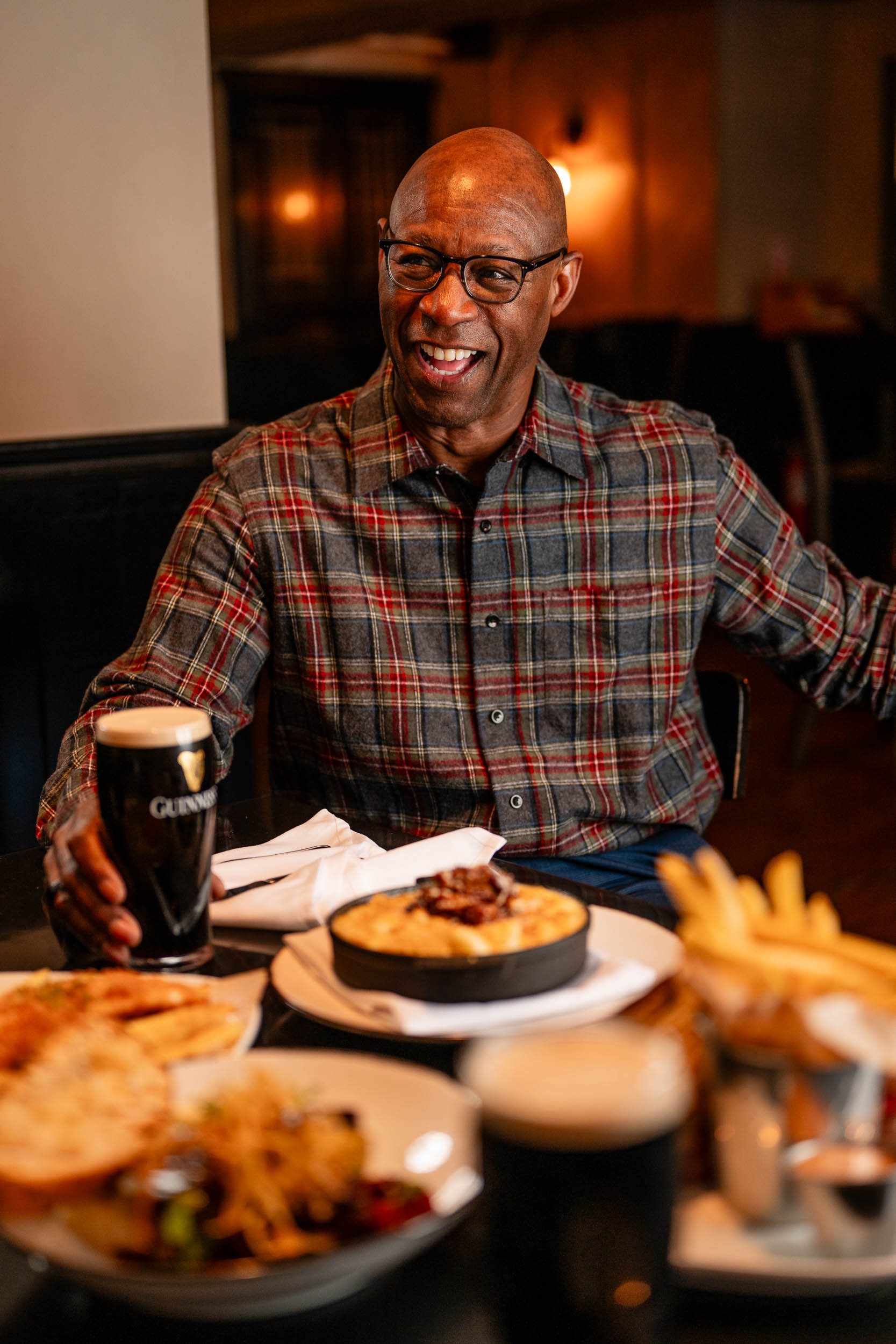 a man wearing a flannel holding a full Guinness smiling at a table with an assortment of food