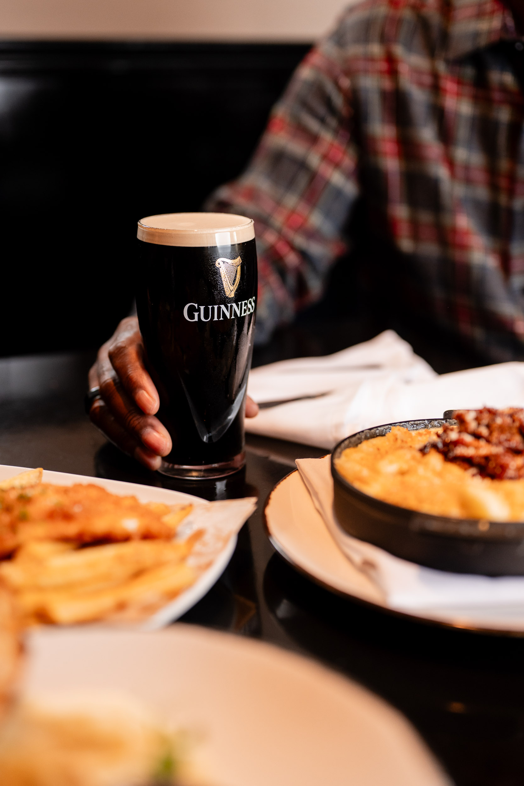 a cold Guinness on a table, held my a man in the background with several dishes in the foreground